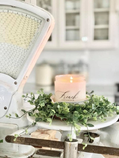 A lit lavender candle on a vintage scale, surrounded by greenery, in a bright kitchen setting.