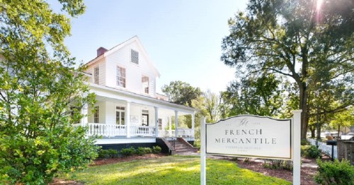 A charming white house with a porch, surrounded by greenery, featuring a sign that reads "French Mercantile."