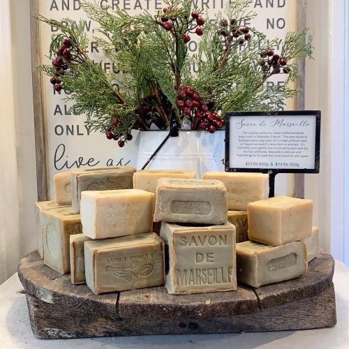 A display of Marseille soap bars on a wooden stand, with greenery and a sign detailing prices in the background.