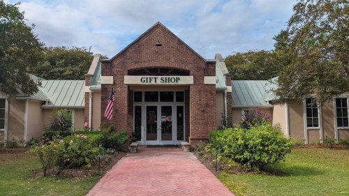 A brick building with a sign reading "GIFT SHOP," surrounded by greenery and an American flag out front.