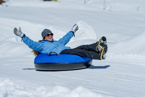 A person in a blue jacket and sunglasses joyfully tubing down a snowy slope, arms raised in excitement.