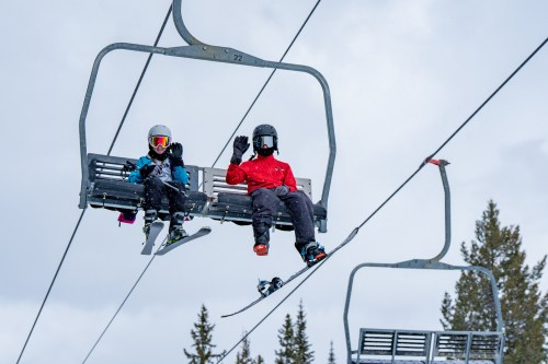 Two children on a ski lift, one in a blue jacket and helmet, the other in red, both waving with snowy trees in the background.