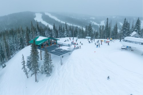 A snowy mountain landscape with a ski lodge, skiers, and tall pine trees under a cloudy sky.