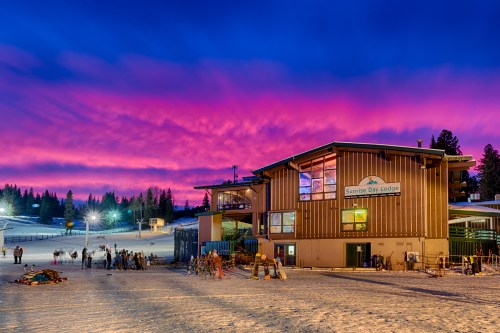 A ski lodge at sunset, with vibrant pink and purple clouds above, surrounded by snow and skiers preparing to hit the slopes.