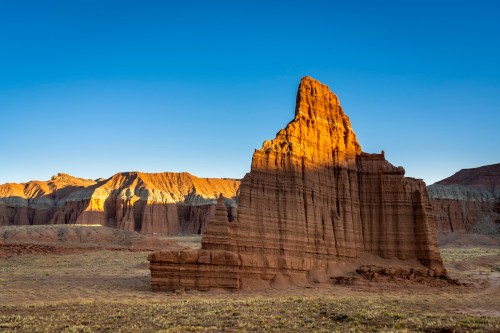 A towering rock formation bathed in golden sunlight, surrounded by rugged terrain and clear blue skies.