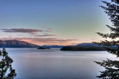 A serene lake at sunset, surrounded by mountains and trees, with soft clouds reflecting on the water's surface.