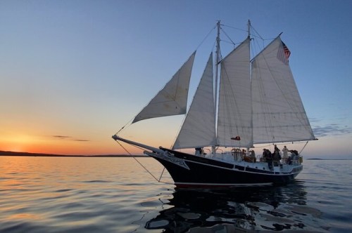 A sailboat with white sails glides across calm waters at sunset, with a colorful sky in the background.