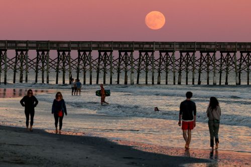 People walking along a beach at sunset, with a large moon rising above a pier in the background.
