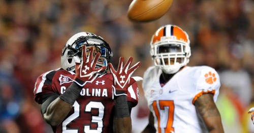 A football player in a maroon jersey attempts to catch a ball, while another player in an orange jersey looks on.