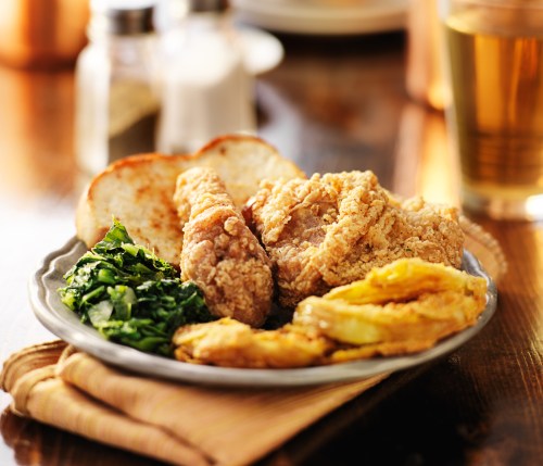 A plate of fried chicken, collard greens, fried plantains, and toasted bread on a wooden table.