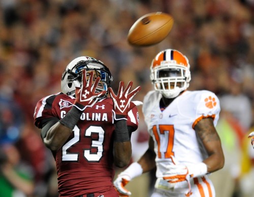 A football player in a maroon uniform attempts to catch a ball, while another player in orange and white looks on.