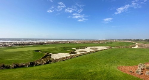 A scenic view of a golf course by the ocean, featuring lush green fairways and sandy bunkers under a clear blue sky.