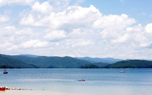 A serene lake surrounded by mountains under a partly cloudy sky, with a small boat and a person kayaking.
