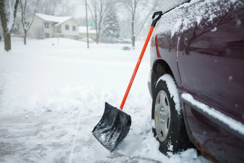 A snow shovel leans against a car in a snowy driveway, with a house visible in the background.