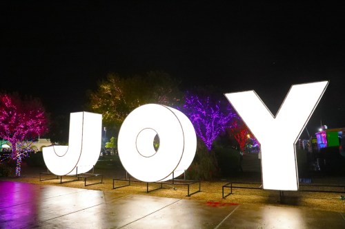 Brightly lit letters spelling "JOY" at night, surrounded by colorful illuminated trees.