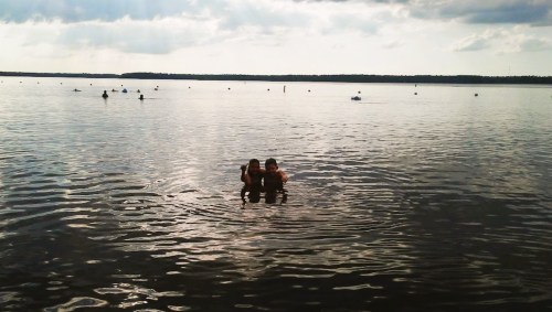 Two people wade in a calm lake, surrounded by a serene landscape and distant boats under a cloudy sky.