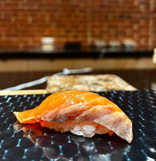 A close-up of a piece of sushi with salmon on a textured black plate, set against a blurred brick background.