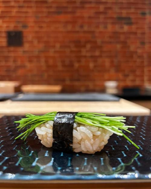 A piece of sushi topped with green chives, resting on a textured black plate against a brick wall background.