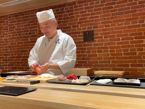 A sushi chef prepares sushi at a wooden counter, with various sushi pieces displayed on trays in front of him.