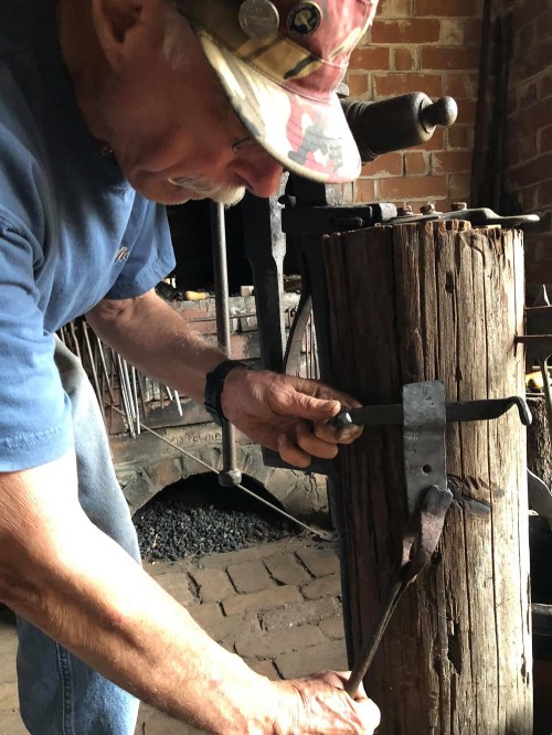 A man in a cap works on a metal piece using tools at a forge, with a wooden post and brick wall in the background.