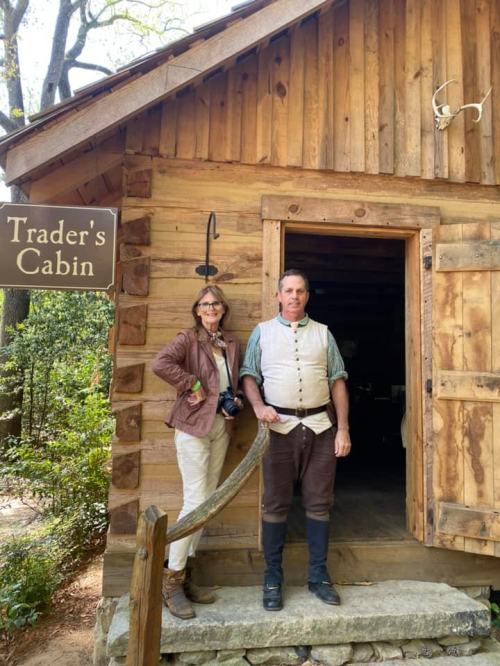 A woman and a man in historical attire stand outside a wooden cabin labeled "Trader's Cabin."