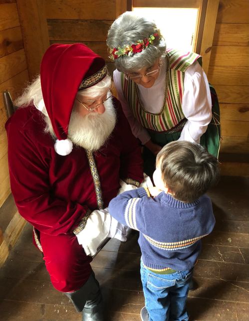 A child interacts with Santa Claus and a woman in traditional attire inside a cozy wooden room.