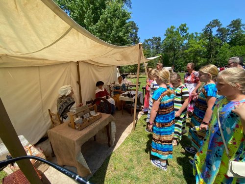 A group of children in colorful dresses watches a demonstration at a historical tent event.
