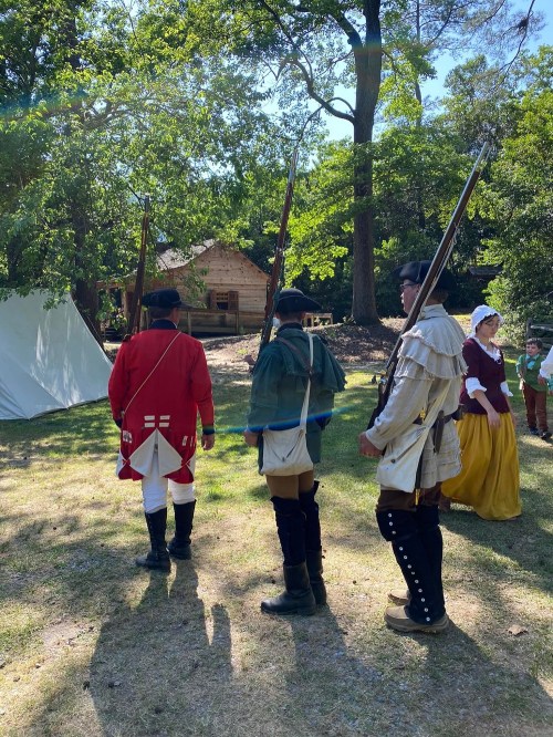 Three historical reenactors in period costumes stand in a grassy area, with a tent and cabin in the background.