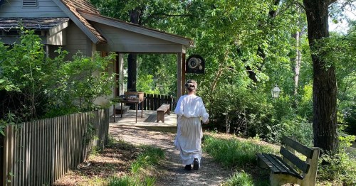 A person in a white dress walks along a path in a lush, green garden near a quaint house.