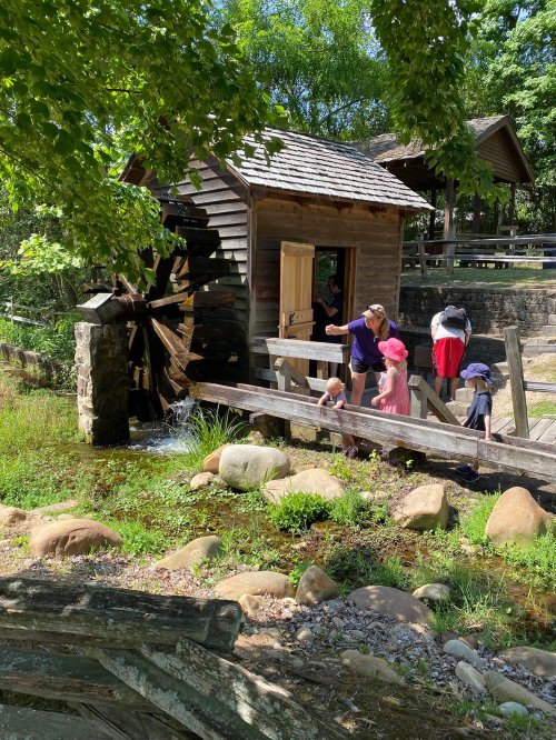 A wooden water mill with a large wheel, surrounded by greenery, as children explore nearby rocks and water.