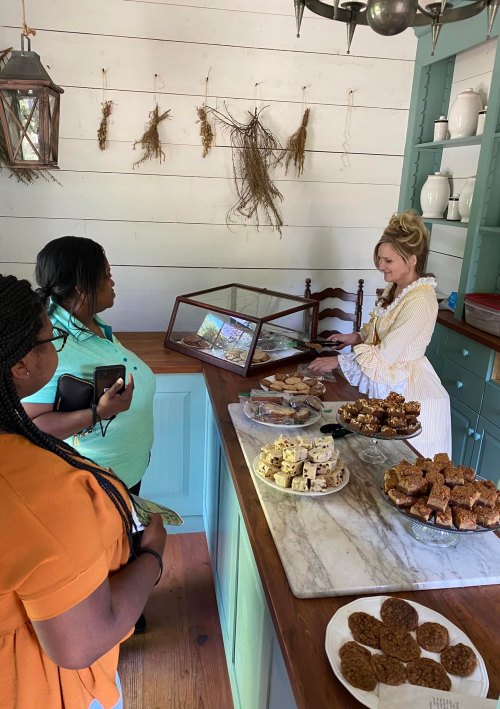 Two women observe a display of baked goods in a historic kitchen, while a woman in period attire serves treats.