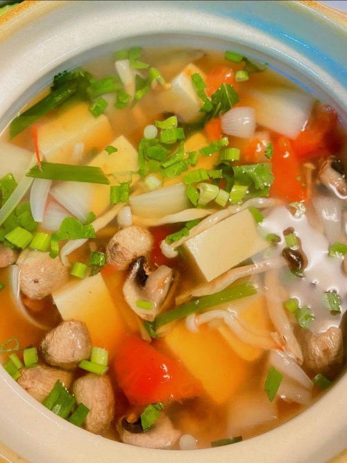 A bowl of colorful vegetable soup with mushrooms, tomatoes, and green onions in a clear broth.