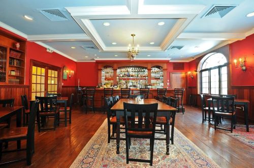 Cozy restaurant interior with wooden tables, chairs, a bar, and red walls, featuring warm lighting and a patterned rug.