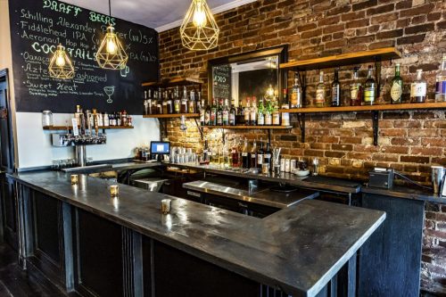 A rustic bar with a brick wall, shelves of liquor, and a chalkboard menu featuring craft beers.