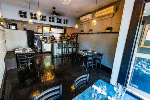 A cozy restaurant interior with wooden tables, black chairs, and a kitchen in the background, featuring warm lighting.