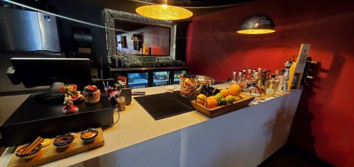 A modern bar counter with a variety of drinks, fruits, and decorative items against a red wall.