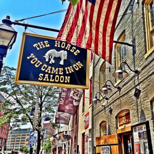 Sign for "The Horse You Came In On Saloon" with an American flag, set in a historic street scene.