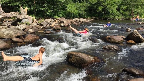 Two children float on inner tubes in a rocky river, enjoying a sunny day while others play in the background.