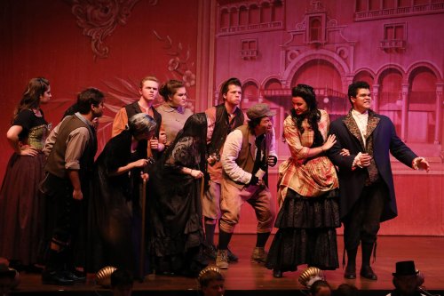A theatrical performance featuring a diverse cast in period costumes, engaged in a lively scene on stage.