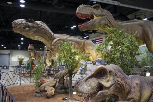 A display of life-sized dinosaur models in a convention center, surrounded by greenery and a U.S. flag in the background.