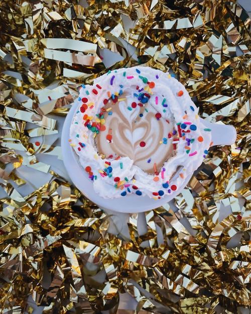 A cup of coffee topped with whipped cream and colorful sprinkles, surrounded by shiny gold confetti.