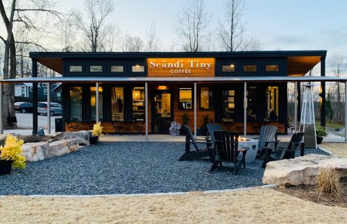 A cozy coffee shop with a wooden exterior, outdoor seating, and a sign reading "Scandi Tiny Coffee" surrounded by trees.