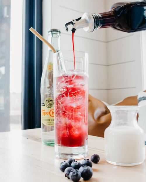 A glass filled with ice and a red drink being poured, alongside a bottle of sparkling water, milk, and blueberries.