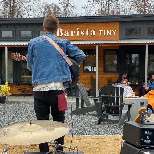 A musician performs with a guitar in front of a café called "Barista Tiny," while people relax nearby.