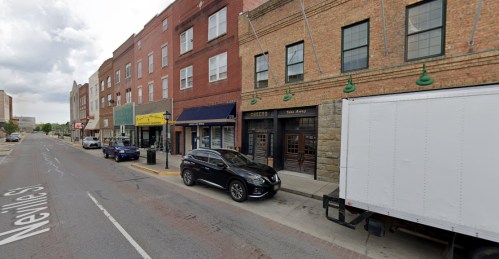 Street view featuring brick buildings, parked cars, and a delivery truck along a city sidewalk.