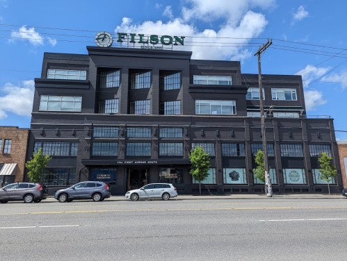 A large black building with "FILSON" sign on top, featuring large windows and trees in front, set against a blue sky.