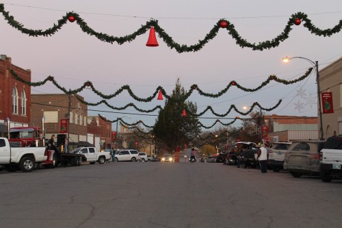 A festive street decorated with garlands and red cones, lined with vehicles and buildings, during twilight.