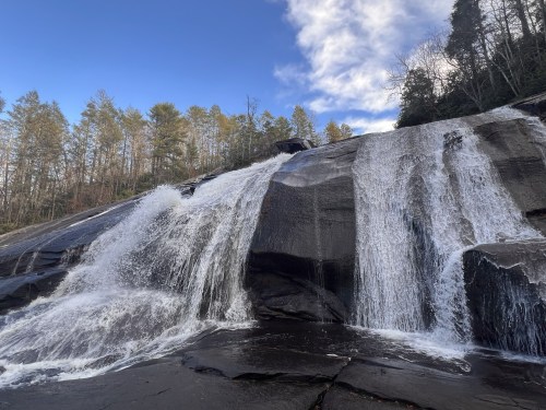 A cascading waterfall flows over dark rocks, surrounded by trees under a blue sky with scattered clouds.