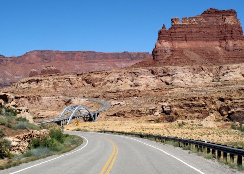 A winding road leads through a desert landscape with red rock formations and a modern bridge in the distance.