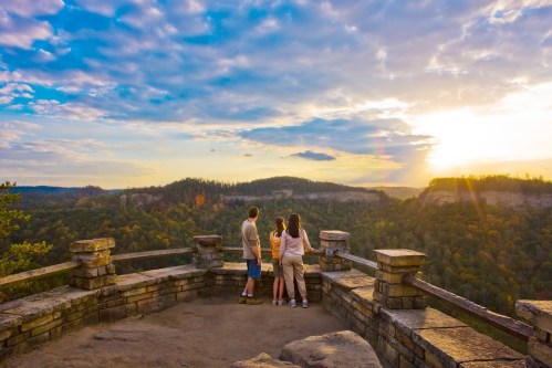 Three people stand on a stone overlook, gazing at a colorful sunset over a lush, mountainous landscape.